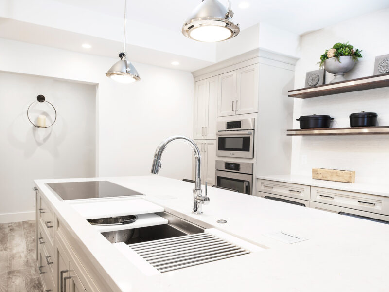 Tulsa kitchen design and remodel featuring a Galley Workstation kitchen sink, island cabinet storage, pendant lights, white cabinets with crown molding, tall storage with Bosch ovens, and floating shelves over wood flooring.