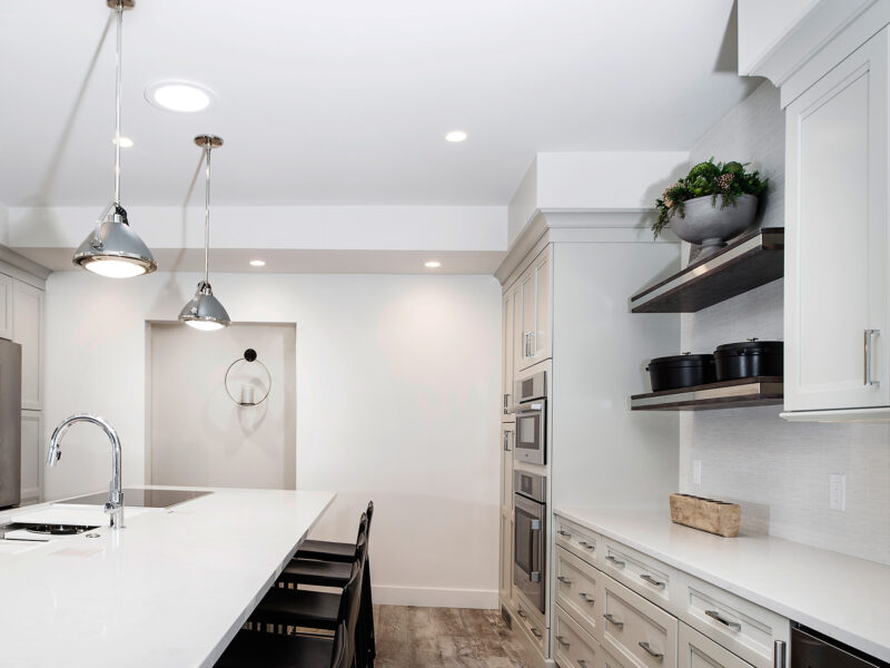 Tulsa kitchen remodel featuring a kitchen island with seating, pendant lights, white cabinet and drawer storage, floating shelves, stainless steel appliances, tall storage, and vinyl wood floors.