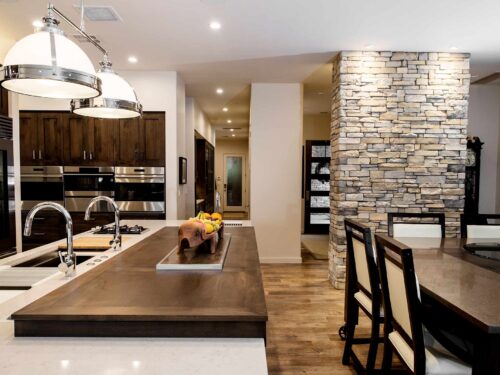 Tulsa kitchen featuring a large island with a Galley Workstation, Wolf two burner gas cooktop, toll away table, seating, a pop-up television, a Caesarstone countertop, a stone wall, decorative pendant lights, and Wolf convection steam ovens.