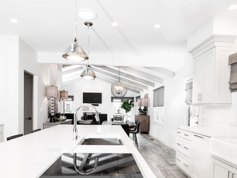 Tulsa kitchen design featuring an open-concept, modern kitchen with a large island, a Galley Workstation, induction cooktop, pendant lights, white cabinet storage, vinyl wood floors, and an adjoining living room with a vaulted ceiling.