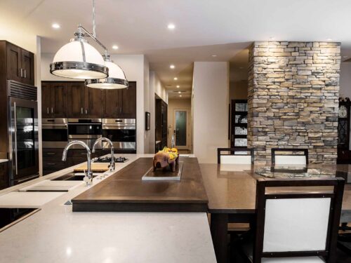 Open Tulsa kitchen featuring a large island with a movable wood table, seating, a pop-up television, a Caesarstone countertop, a stone wall, decorative pendant lights, and Wolf convection steam ovens.
