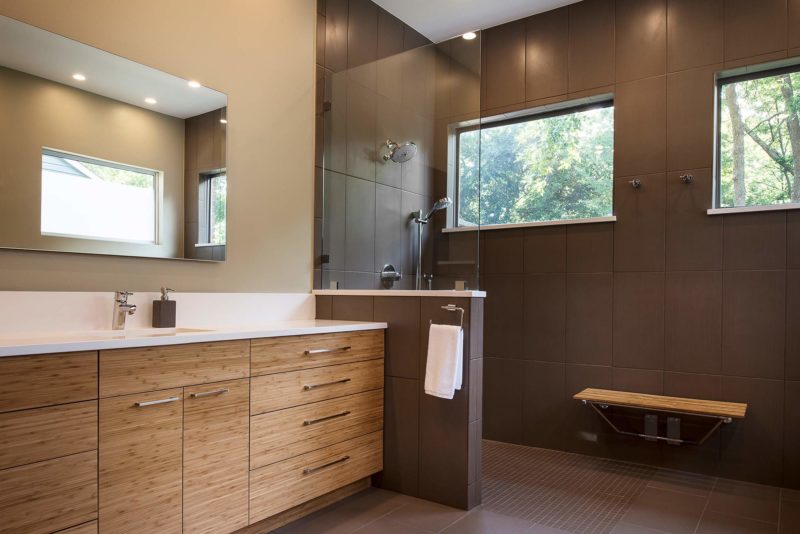 Modern Tulsa designed and remodeled master bathroom with medium brown wood grain base cabinet storage, walk-in shower glass partition, large wall tile and shower bench