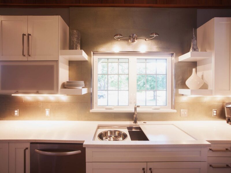 Tulsa kitchen remodel featuring a dishwasher, a sink base storage, a Galley Workstation kitchen sink, white counters, a tile backsplash, floating shelves, and a sconce light.