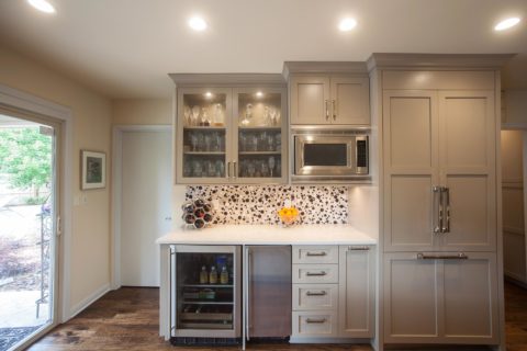Tulsa kitchen remodel featuring a white kitchen beverage space with a microwave, an undercounter refrigerator and ice maker, a tile backsplash, wall cabinets with beaded glass doors, and a paneled refrigerator.