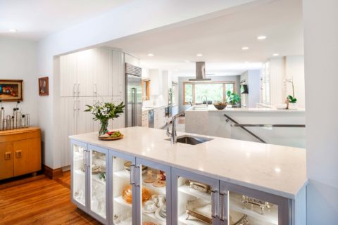 Tulsa kitchen design and remodel featuring an open white kitchen with marble countertops, Bosch professional stainless steel appliances, a lit, glass-front bar island with storage and a bar sink, all on wood floors.