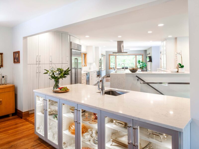 Tulsa kitchen design and remodel featuring an open white kitchen with marble countertops, Bosch professional stainless steel appliances, a lit, glass-front bar island with storage and a bar sink, all on wood floors.