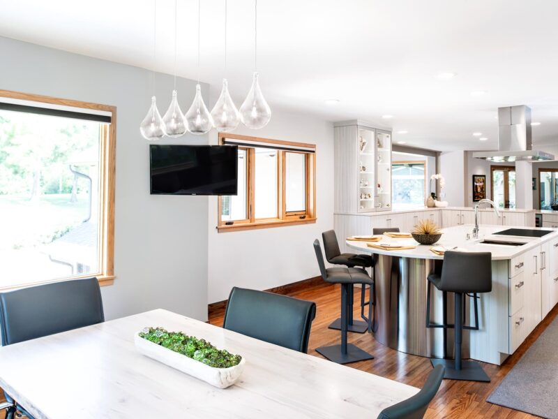 Tulsa kitchen remodel featuring a dining table island with an induction cooktop, a Galley Workstation, and a Bosch island vent hood. The space includes white counters, seating, tall wall cabinets with glass panel doors, and wood floors.