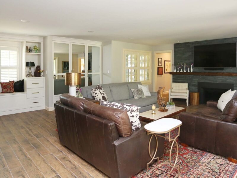 Modern open-concept living room featuring staggered gray stone fireplace with wood mantle, large wall-mounted TV, white cabinet storage, and rich brown wood plank flooring in Tulsa remodel.