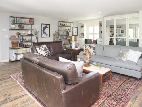 Open living room featuring comfortable sofa seating, decorative wall shelves, flush tall white cabinet storage, and rich medium brown wood flooring in Tulsa kitchen remodel.