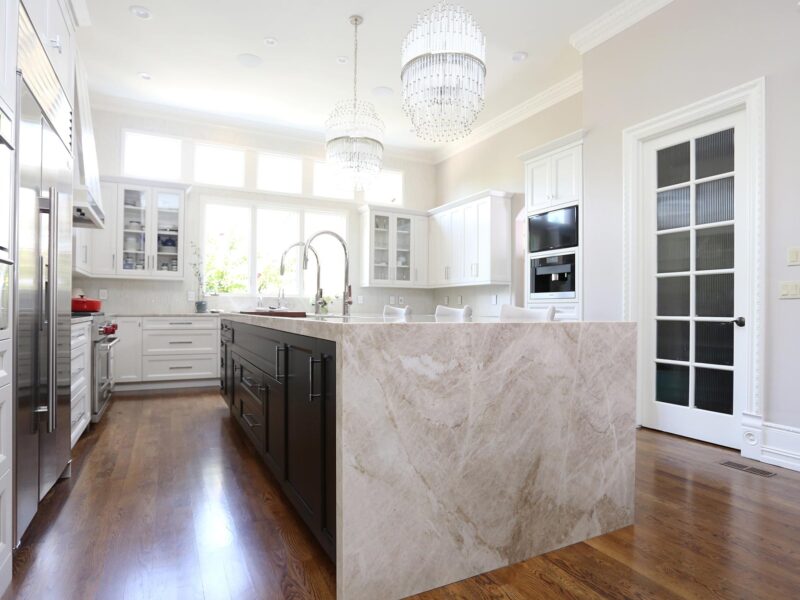 Tulsa kitchen design and remodel featuring a large kitchen island with brown cabinet storage and a waterfall edge countertop. The space includes white perimeter cabinets, refinished wood floors, Sub-Zero and Wolf stainless steel appliances, and a Galley Workstation.