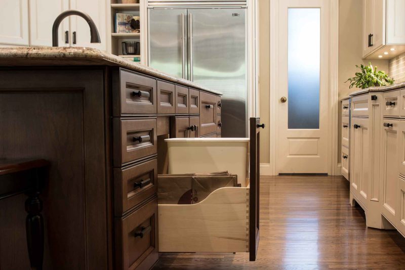 Kitchen island pullout trash, brown cabinet drawer storage, cream base cabinet storage, wood flooring, Sub-Zero freezer refrigerator Tulsa kitchen design and remodel