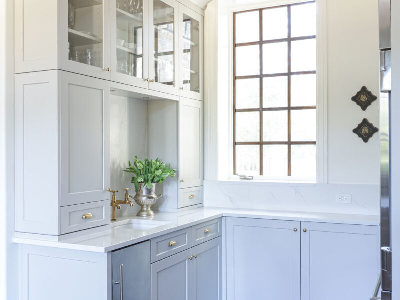 Tulsa kitchen remodel featuring a beverage bar with wood floors, an undercounter ice maker, a bar sink, and white cabinets with glass fronts and crown molding.