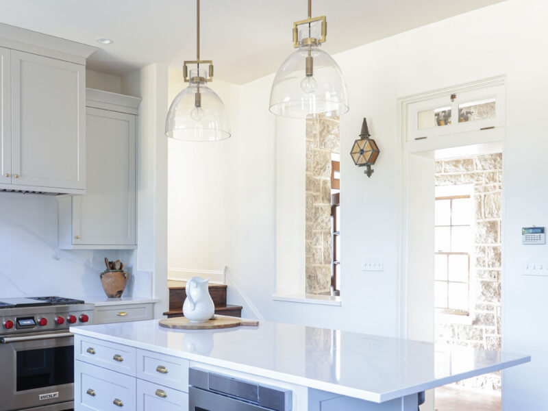 Tulsa kitchen remodel featuring a kitchen island with a built-in drawer microwave and pullout trash. The space has white kitchen cabinet storage, wood floors, and is illuminated by pendant lights over a Wolf gas range.