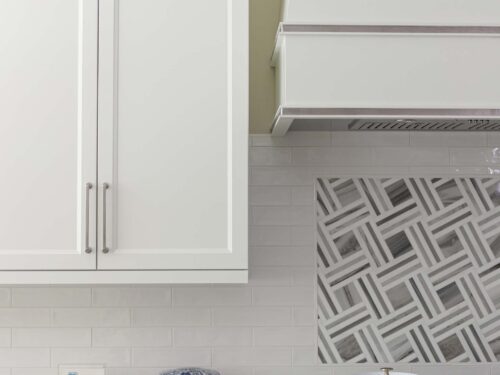 Tulsa kitchen design and remodel featuring a range top, subway tile backsplash, a decorative vent hood, and white cabinet storage.