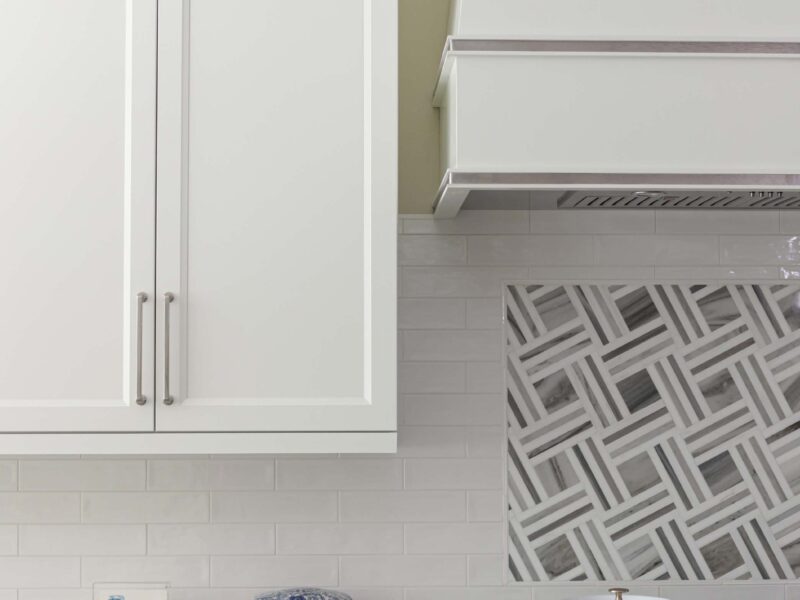 Tulsa kitchen design and remodel featuring a range top, subway tile backsplash, a decorative vent hood, and white cabinet storage.