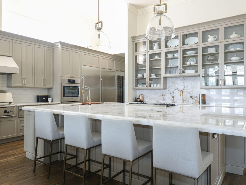 Tulsa kitchen designer and remodeler showcasing a large kitchen island with seating, a built-in oven, and a cooking area. The space features pendant lights, a full-size refrigerator and freezer, and a tall wine cabinet with storage.