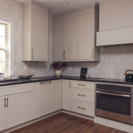 Greige kitchen cabinet with a built-in oven and black counters. Features include a workstation sink, a tile backsplash, and smart storage.