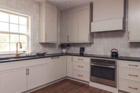 Greige kitchen cabinet with a built-in oven and black counters. Features include a workstation sink, a tile backsplash, and smart storage.