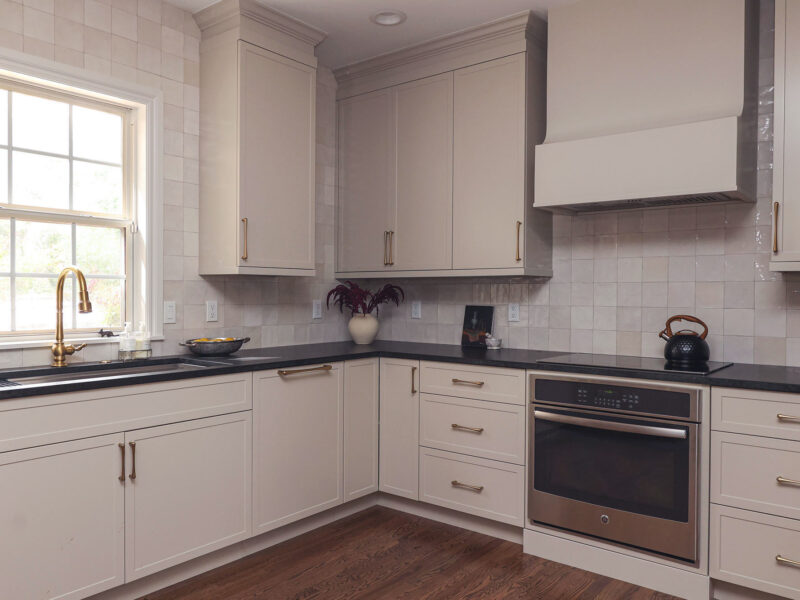 Greige kitchen cabinet with a built-in oven and black counters. Features include a workstation sink, a tile backsplash, and smart storage.